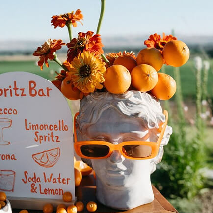 Buste antique coiffé d'oranges et de fleurs, lunettes orange, devant l'ardoise d'un bar à spritz
